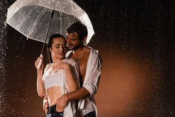 Wall Mural - smiling man holding hands in pockets of sexy woman standing under rain with transparent umbrella on dark background