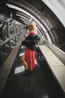 © Vimaki - Mujer joven posando en pasillo metro de barcelona con luz artificial interior, vestido rojo, chaqueta de cuero, gafas rubia, botas negras y pierda.