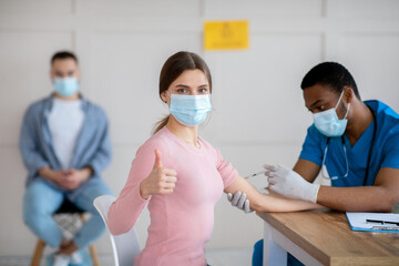 Poster - Young woman in medical mask approving of covid-19 vaccination, showing thumb up gesture at clinic