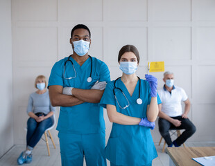 Poster - Male and female doctors wearing uniforms and face masks, holding syringe with coronavirus vaccine at clinic