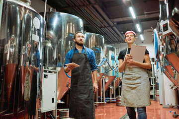 Two young workers of large beer production factory discussing new equipment