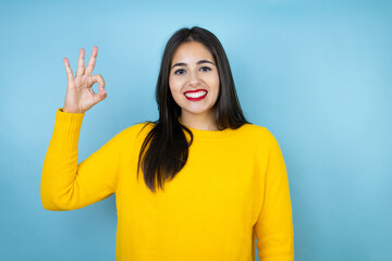 Young beautiful woman wearing yellow sweater over isolated blue background doing ok sign with fingers and smiling, excellent symbol