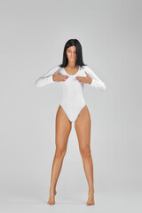 Wall Mural - beautiful slim tanned woman with perfect body and long legs in white bodysuit posing over studio background.