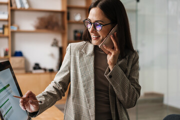 Wall Mural - xSmiling woman talking on cellphone and working with computer in office