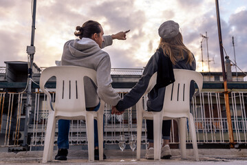 Lovely couple sitting on the chairs at rooftop holding hands and looking to the sky.