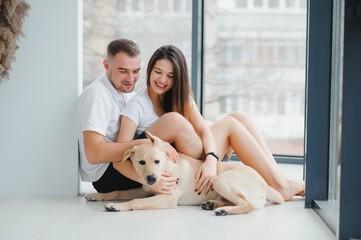 The young happy couple is moving into a new house. They are sitting down on the floor with their little puppy after they brought boxes with things to their new home.