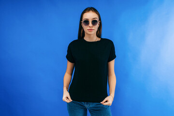 Wall Mural - Stylish girl in glasses wearing black t-shirt posing in studio on blue background