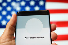 Close Up An Adult Hand Holding A Smartphone Showing "Account Suspended" From A Social Media Application Against The American Flag Background. 