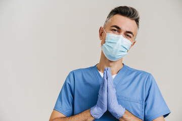 Pleased medical doctor in face mask and gloves holding palms together