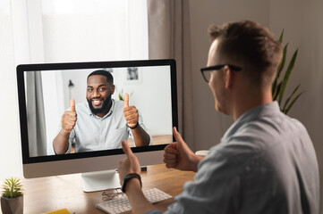 Video call, video meeting. A young man is discussing some business tasks with a young African man via video, they have come to a consensus and show thumbs up as a sign of successful negotiations