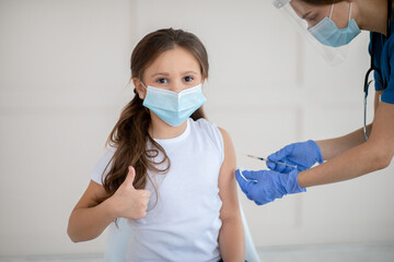 Poster - Little girl in face mask gesturing thumb up while receiving covid-19 vaccine injection at health centre
