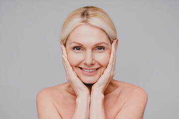 Nice-looking middle-aged caucasian woman smiling and touching her fresh clean face isolated over grey background