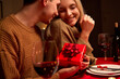 © insta_photos - Happy young couple celebrating anniversary or Valentines day having romantic dinner at home table. Loving husband giving red gift box hugging wife making present surprise on date in candle light.