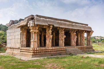 Hampi, Karnataka, India - November 5, 2013: KudureGombe mandapam and ancient well. Closeup of the brown stone building set in green environment under blue cloudscape. Horse sculptures.