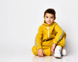 © FAB.1 - Schoolboy dressed in a stylish yellow sports suit and white sneakers poses on a white background. Child looks into the camera sitting near a free space for text. Children's sports fashion concept.