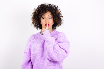 Surprised young beautiful caucasian woman wearing pink knitted sweater against white wall makes silence gesture, keeps finger over lips and looks mysterious at camera