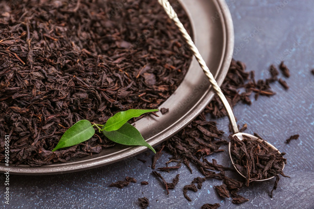 Dry black tea leaves in plate with spoon on dark background