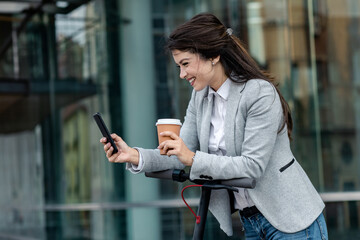 Wall Mural - Businesswoman with electric scooter standing in front of modern business building looking at phone.