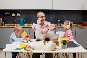 Father with a newborn baby in arms working from home during quarantine and closed school. coronavirus outbreak. Young businessman freelancer works on laptop with children playing around.