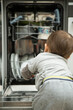 © Dina - Little child boy toddler helping mom to disassemble the dishwasher. The dishwasher is open, the child is sitting next to him and holding clean dishes in his hands.