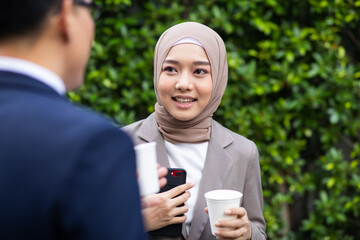 Wall Mural - Young business muslim woman and business man colleague walking and discussing outside the office. Woman is holding a coffee and businessman is holding a digital tablet outdoor.