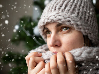 A beautiful young woman in a warm knitted hat and scarf watches the snow fly.