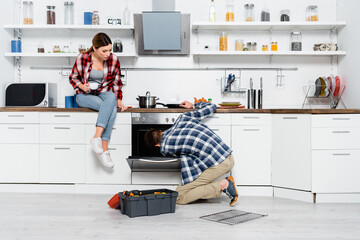 Wall Mural - full length of young woman with coffee cup sitting on table near man repairing oven in kitchen