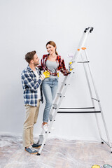 Canvas Print - full length of smiling young man hugging woman with paint roller standing on ladder at home