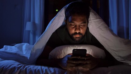 Poster - technology, internet, communication and people concept - young indian man with smartphone lying in bed under blanket at home at night