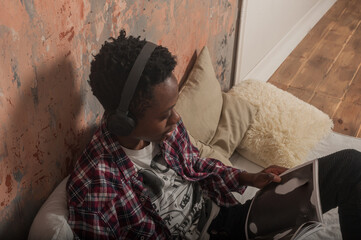 a young african american man lies on the bed wearing headphones and reads a magazine