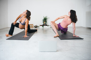 Portrait of two young millennial women friends make yoga exercises watching a tutorial on laptop - Pregnant people performs workouts with teacher