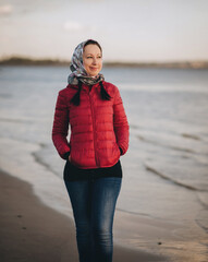 A young European girl in a headscarf and a red jacket stands on the bank of a river (lake) and looks to the side. The concept of an autumn (spring) walk in the evening sunset time.