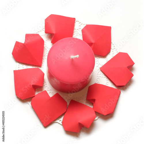 Thick pink aromatic candle on white openwork paper napkin, surrounded by a several 3D handmade paper hearts on white background top closeup view isolated. Love, rendezvous, relations, dating concept 