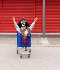 two white caucasian men playing riding in a shopping cart jumping in a parking lot happy and happy enjoying they go shopping with red and blue background