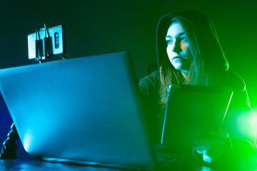Poster - Freelancer girl next to a laptop. Girl is illuminated by a neon light. Woman freelancer during work. Laptop and two phones next to freelancer. Concept - it works remotely. Woman works with a computer