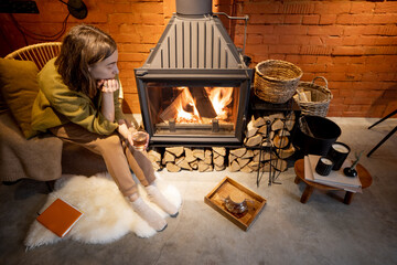 Young thoughtful woman sitting by a burning fireplace, looking on the flame in cozy house