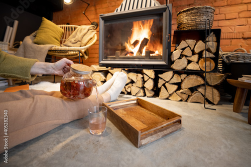 Obraz Dibond Woman lying with a hot drink by the burning fireplace in a cozy house in loft style