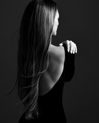 Young pretty woman model standing backwards in dress with open back and showing her gorgeous silky long straight hair over dark background. Hairstyle, hairsalon, hairdresser, fashion concept