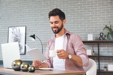 Young man with laptop reading magazine and drinking tea at home