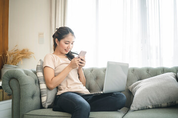 Happy casual beautiful Asian woman using mobile phone while sitting on the couch in the house.