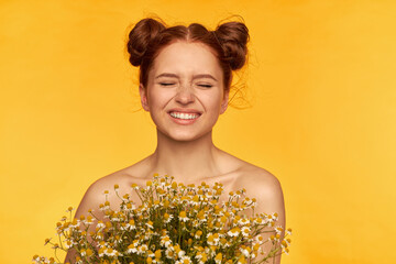 Portrait of attractive, cute, charming, red hair girl with buns. Holding a bouquet of wildflowers and squints in a smile. Healthy skin. Closeup, stand isolated over yellow background