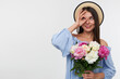 © timtimphoto - Portrait of attractive girl with long brunette hair. Wearing a hat and blue dress. Holding a bouquet of beautiful flowers and watch through fingers to the left at copy space over white background