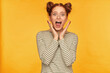 Young lady, horrified looking ginger woman with two buns. Shows how much she frightened of what she see. Wearing striped sweater and screams at the camera isolated over yellow background