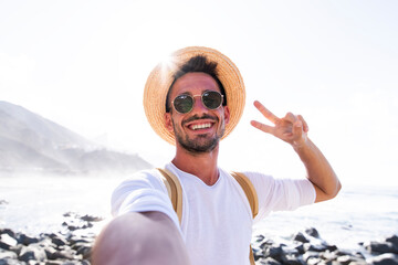 Wall Mural - Happy young man with hat and backpack taking a selfie portrait hiking on mountains - Pov view of a smiling guy using smart phone mobile 