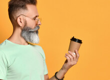 Brutal Elderly Man With A Beard Drinking Takeaway Coffee In The Paper Cup On A Yellow Background With A Serious Face, Looking At The Glass
