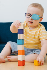 little blond toddler boy in a lazy eye patch is playing with toy cube at home