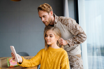 Wall Mural - Military man doing hair of his daughter while she using tablet computer