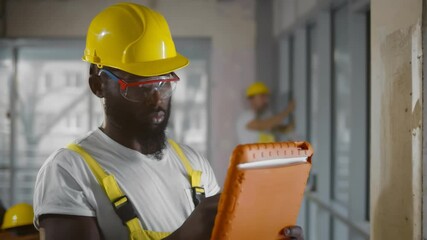 Wall Mural - Afro-american builder in safety glasses and hardhat using tablet computer at construction site