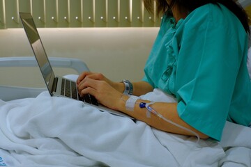 A half body shot of a workaholic business woman sitting on her hospital bed with IV injection in her arm, recovering from surgery while using her laptop computer to continue her work.