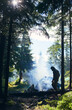 © anatoliy_gleb - Guy in jacket and hood making fire and smoke flows up. Tent among fir trees under the rays of sun breaking through clouds on cold day. Camping in the forest